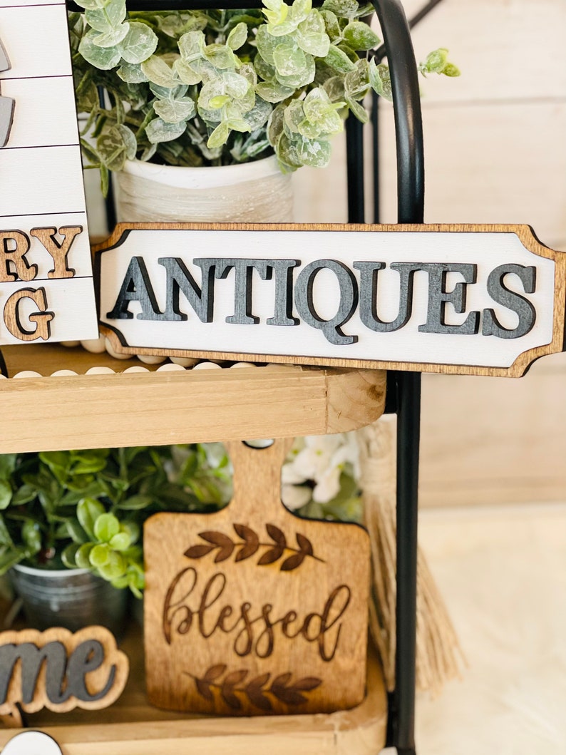 May include: A wooden sign with the word "ANTIQUE" in black letters on a white background. The sign is on a tiered wooden shelf with greenery and other decorative items.
