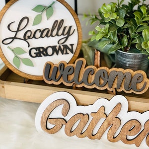 May include: Three wooden signs with white backgrounds and black lettering. The first sign is round and says "Locally Grown" with green leaves around the text. The second sign says "welcome" and the third sign says "gather".
