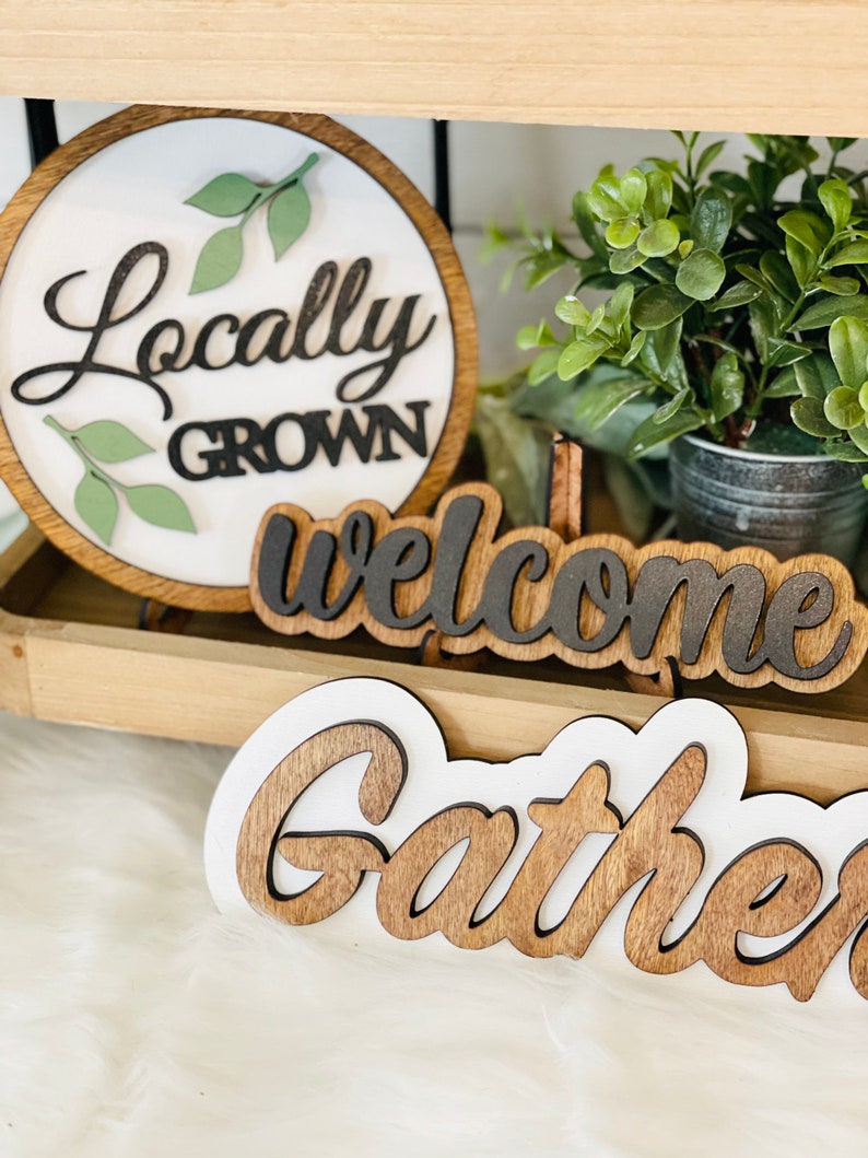 May include: Three wooden signs with white backgrounds and black lettering. The first sign is round and says "Locally Grown" with green leaves around the text. The second sign says "welcome" and the third sign says "gather".