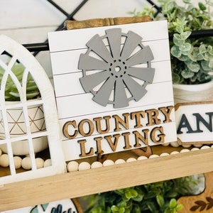 May include: A wooden sign with a gray windmill cutout and the words "COUNTRY LIVING" in brown letters. The sign is on a shelf with other farmhouse decor, including a white planter with a succulent, a white planter with eucalyptus, and a wooden sign with the word "AND" in black letters.