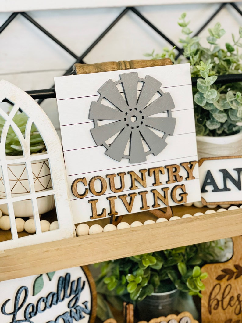 May include: A wooden sign with a gray windmill cutout and the words "COUNTRY LIVING" in brown letters. The sign is on a shelf with other farmhouse decor, including a white planter with a succulent, a white planter with eucalyptus, and a wooden sign with the word "AND" in black letters.