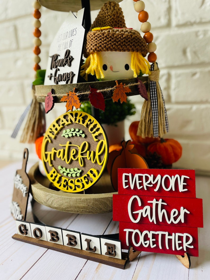 Może przedstawiać: Tr&oacute;jpoziomowa taca ozdobiona na jesień z postacią straszakiem, dyniami i tabliczkami z napisami "Let our lives be full of thanks + giving", "Grateful Blessed" i "Everyone Gather Together".