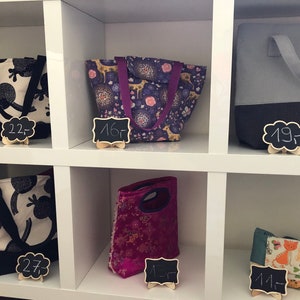 Puede incluir: Seis bolsas de mano y bolsas de almuerzo diferentes que se muestran en estantes blancos. Las bolsas están hechas de tela y tienen varios patrones y colores, incluyendo blanco y negro, morado y rosa, y azul y gris. Cada bolsa tiene una etiqueta de precio adjunta, que van desde 14 a 27 euros.