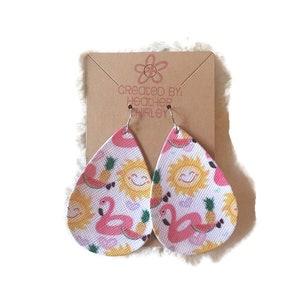 May include: A pair of teardrop-shaped earrings with a summer theme. The earrings feature a repeating pattern of pink flamingos, smiling suns, pineapples, and watermelon slices on a white background. The text "created by: Heather Shirley" is visible on the backing card.
