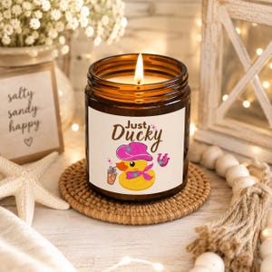 Op de afbeelding: Een bruine glazen kaars met een wit etiket met een gele badeend met een roze hoed en sjaal, die een drankje vasthoudt. De tekst luidt Just Ducky. De kaars brandt en staat op een ronde gevlochten onderzetter, omringd door een witte zeester.