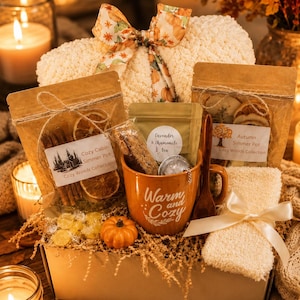 May include: A gift box containing a cream-colored plush blanket with a pumpkin-print ribbon, two pouches of simmer pot ingredients, a bag of lavender and chamomile tea, a metal tea infuser, a wooden spoon, and an orange mug labeled Warm and Cozy.