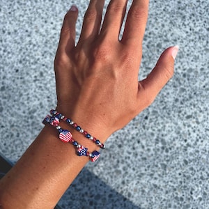 May include: Two bracelets with patriotic themes are worn on a wrist. One bracelet features red, white, and blue beads with star and flag charms. The other bracelet has small, dark beads. The bracelets are set against a textured, light gray background.