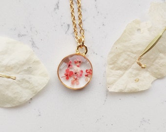 May include: A gold chain necklace with a round pendant featuring pressed pink flowers encased in clear resin.