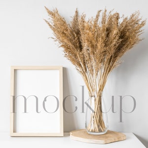 May include: A wooden frame with a white background sits on a white surface next to a vase of dried pampas grass. The vase is clear glass and sits on a wooden base.