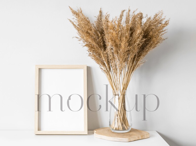 May include: A wooden frame with a white background sits on a white surface next to a vase of dried pampas grass. The vase is clear glass and sits on a wooden base.