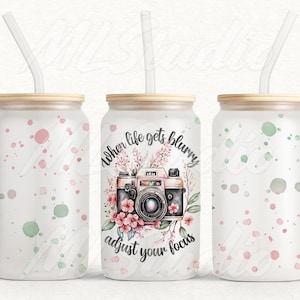 May include: Three white stainless steel tumblers with a wood lid and a clear plastic straw. The centre tumbler has a pink floral design with a vintage camera and the text "When life gets blurry adjust your focus". The tumblers have a white background with pink and green watercolour dots.