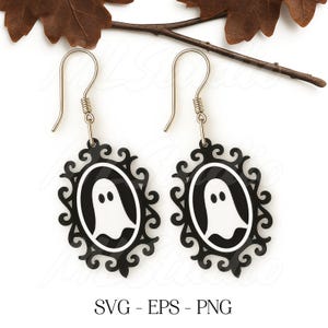 Op de afbeelding: Een paar Halloween-oorbellen. Elke oorbel heeft een zwart, sierlijk ovaal frame met een wit spooksilhouet. De oorbellen hangen aan zilverkleurige oorhaakjes. De tekst "SVG - EPS - PNG" staat onderaan.