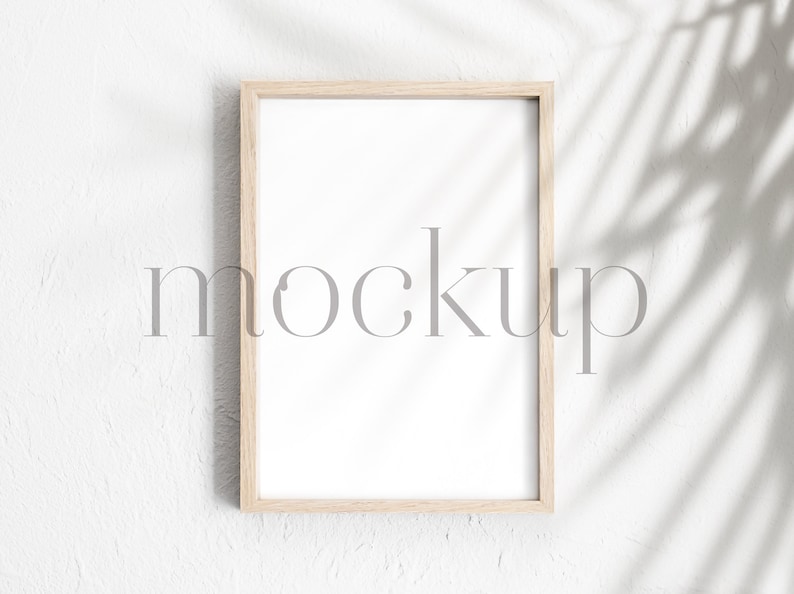 May include: A light wood frame with a white mat and a blank white space inside. The frame is hanging on a white wall with a shadow of a plant leaf behind it. The word "mockup" is written in gray text in the center of the image.