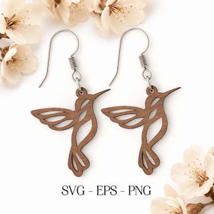 May include: A pair of wooden hummingbird earrings with silver-colored ear wires. The earrings are a light brown color and feature a cutout design of a hummingbird. The text "SVG - EPS - PNG" is printed at the bottom of the image.