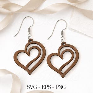 以下が含まれることがあります： 切り絵のデザインが施された、木製のハート型イヤリングのペア。イヤリングは温かみのある茶色で、銀色のイヤーフックから吊り下げられています。画像には「SVG - EPS - PNG」の文字が含まれています。