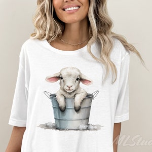 Puede incluir: Camiseta blanca con un gr&aacute;fico de un cordero blanco sentado en un cubo de metal gris. El cordero tiene orejas rosas y una expresi&oacute;n dulce.