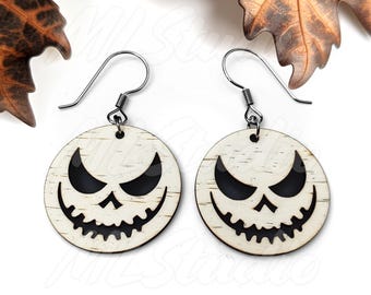 May include: A pair of wooden earrings with a white background and a black jack-o'-lantern face cutout. The earrings are attached to silver hooks.
