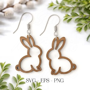 May include: A pair of wooden bunny-shaped earrings with silver-colored hooks. The earrings are a light brown color and feature a cutout design of a bunny rabbit. The text "SVG - EPS - PNG" is printed at the bottom of the image.