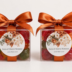 May include: Two clear square favor boxes filled with colorful gummy candies, each topped with a burnt orange satin ribbon bow. A circular white label on the front features a floral illustration and the text: Safa & Hafiz Nikkah 30th May 2026.
