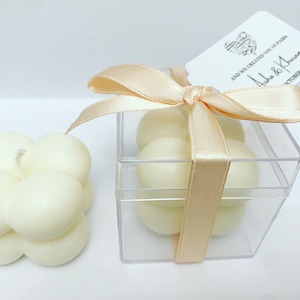 May include: A clear plastic box with a white ribbon tied in a bow. The box contains four small white candles. The box has a white tag attached to it that says "And we created you in Paris" with the names "Luke & Kirsten" and the date "October 2023".
