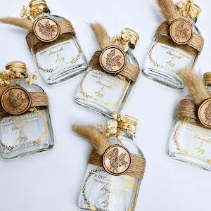 May include: Six small, clear glass bottles with gold lids, each adorned with a wooden leaf-shaped seal and wrapped in twine. The bottles are decorated with dried flowers and feature gold lettering.