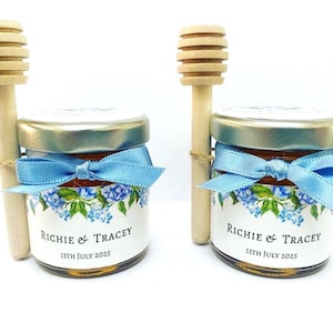 Può includere: Due piccoli vasetti di vetro riempiti di miele, ciascuno con coperchio dorato, nastro azzurro e un bastoncino spargimiele in legno. Le etichette presentano un motivo floreale blu e il testo "Richie & Tracey" seguito dalla data "13th July 2025".