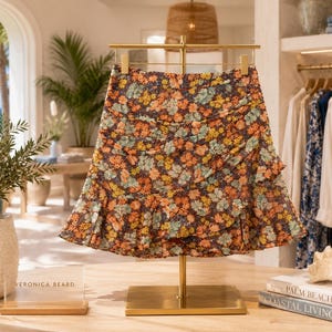 May include: A floral print mini skirt featuring a dark background with orange, yellow, and light green flower patterns. The skirt has a gathered, ruched side detail and a ruffled hemline, displayed on a gold metal stand with a Veronica Beard label.