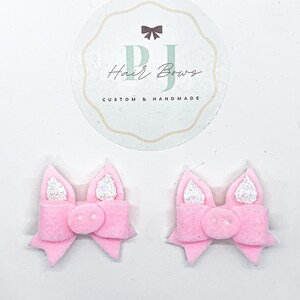 May include: Two pink pig hair bows with white glitter ears. The bows are handmade and have a pig nose in the center.