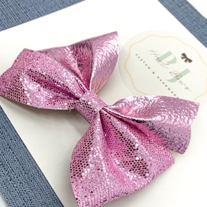 May include: A pink glitter bow with a textured finish. The bow is tied in a classic shape with two loops and a center knot. The bow is on a white background with a small circle sticker that says "PJ's Custom & Handmade".