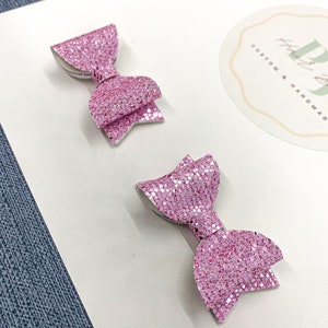 May include: Two pink glitter hair bows on a white background. The bows are small and have a simple design. The background has a sticker with the text "Hutt & Co. Custom & Handmade".