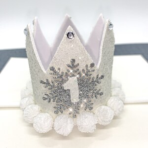 May include: White felt crown with silver glitter and a snowflake design. The crown has a white felt number one in the center of the snowflake and white pom poms around the base.
