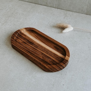 Puede incluir: Una bandeja de madera pequeña de forma ovalada con veta de madera marrón oscuro. La bandeja está hecha de madera natural y tiene un acabado liso y pulido.