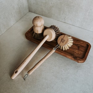 Peut inclure: Trois brosses à vaisselle en bois avec des poils naturels sur un plateau en bois. Les brosses ont des manches en bois et sont étiquetées "TEA" et "DE LUXE" en lettres noires.