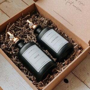 May include: Two black glass bottles with gold spray tops and white labels. The labels read "HUILE" and "VINAIGRE". The bottles are in a brown cardboard box with shredded paper.