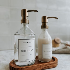 Może przedstawiać: Dwie przezroczyste szklane butelki z dozownikami z pompką w kolorze brązu, z etykietami "Dish Soap" i "Hand Wash", stoją na drewnianej tacy. Butelki są wypełnione przezroczystymi i nieprzezroczystymi płynami. Etykiety są białe z czarnym tekstem. Taca jest owalna.