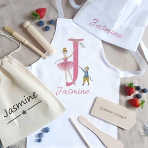 May include: A white apron, chef's hat, and drawstring bag with the name "Jasmine" printed in pink. The apron features a ballerina and a mouse, and the letter "J". Wooden rolling pin, brush, and cutting board are also included.