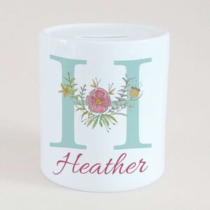 May include: White ceramic money box featuring a light blue letter "H" and the name "Heather" in a cursive font. The letter is decorated with a floral wreath in pink, yellow, and green.