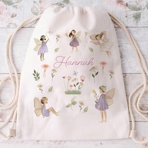 May include: A cream-coloured drawstring backpack with illustrations of fairies, flowers, and mushrooms. The name "Hannah" is written in cursive. The bag has a beige rope drawstring.