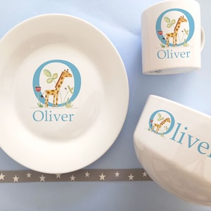 May include: A white ceramic children's dinnerware set with a blue and yellow giraffe illustration. The set includes a plate, bowl, and mug. Each piece features the name "Oliver" in blue lettering.