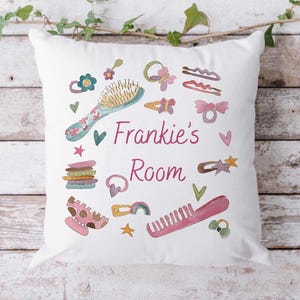 Può includere: Un cuscino quadrato bianco con la scritta Frankie's Room in corsivo rosa. Il design presenta illustrazioni colorate di accessori per capelli come una spazzola blu, un pettine rosa, fermagli, fiocchi ed elastici per capelli.