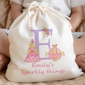 Può includere: Una borsa di tela bianca con coulisse, una lettera E viola, un'illustrazione di una principessa e una carrozza rosa e il testo "Emily's Sparkly Things".