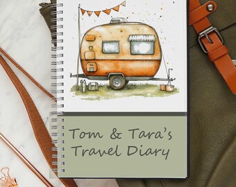 May include: A spiral-bound notebook with a watercolor illustration of an orange camper with a banner. The notebook cover has the text "Tom & Tara's Travel Diary" in a handwritten style. The notebook is next to a brown leather strap and a green bag.