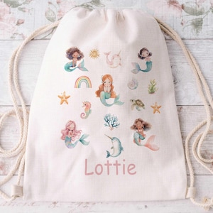 May include: A cream-coloured drawstring backpack with watercolour illustrations of mermaids, a rainbow, starfish, and other sea creatures. The name "Lottie" is printed in pink script on the front. The bag has cream-coloured rope straps.