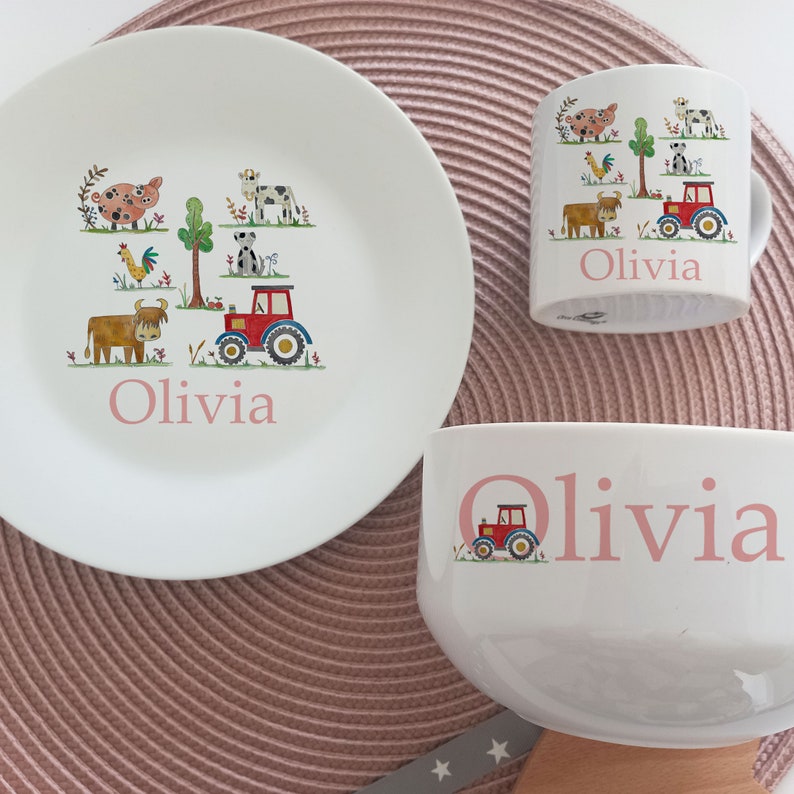 May include: A white ceramic children's dinnerware set with a farm theme. The set includes a plate, bowl, and mug. Each piece features a red tractor, cows, pigs, and a rooster. The name "Olivia" is written in pink on each piece.