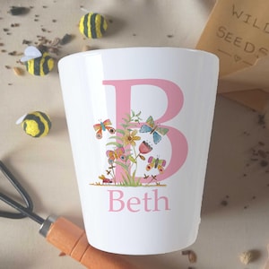 May include: A white ceramic mug with a pink letter B decorated with colourful flowers and butterflies. The name Beth is written below the letter.