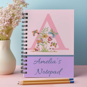 May include: A pink notebook with a large pink letter A decorated with butterflies and flowers. The notebook has a purple band with the text "Amelia's Notepad" in blue.