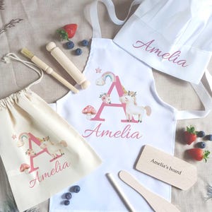 May include: A children's baking set with a unicorn design. The set includes a white apron, chef's hat, drawstring bag, wooden utensils, and a cutting board. The apron and bag feature a pink letter "A" and the name "Amelia". Strawberries and blueberries are also visible.