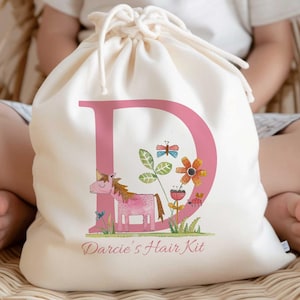 May include: A white cotton drawstring bag with a pink letter D, a unicorn, flowers, and a bee. The bag says "Darcie's Hair Kit" in pink lettering.