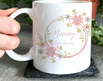 May include: White ceramic mug with a pink floral wreath design. The wreath has pink and yellow flowers and green leaves. The text "Mummy Love Amelia" is inside the wreath.