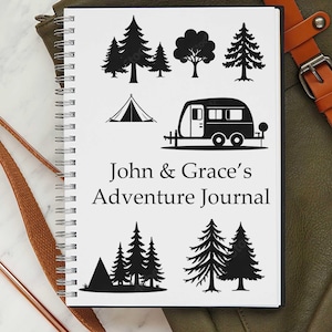 May include: A white spiral-bound notebook with black illustrations of trees, a tent, and a caravan. The text "John & Grace's Adventure Journal" is printed on the cover. The notebook is on a green surface.